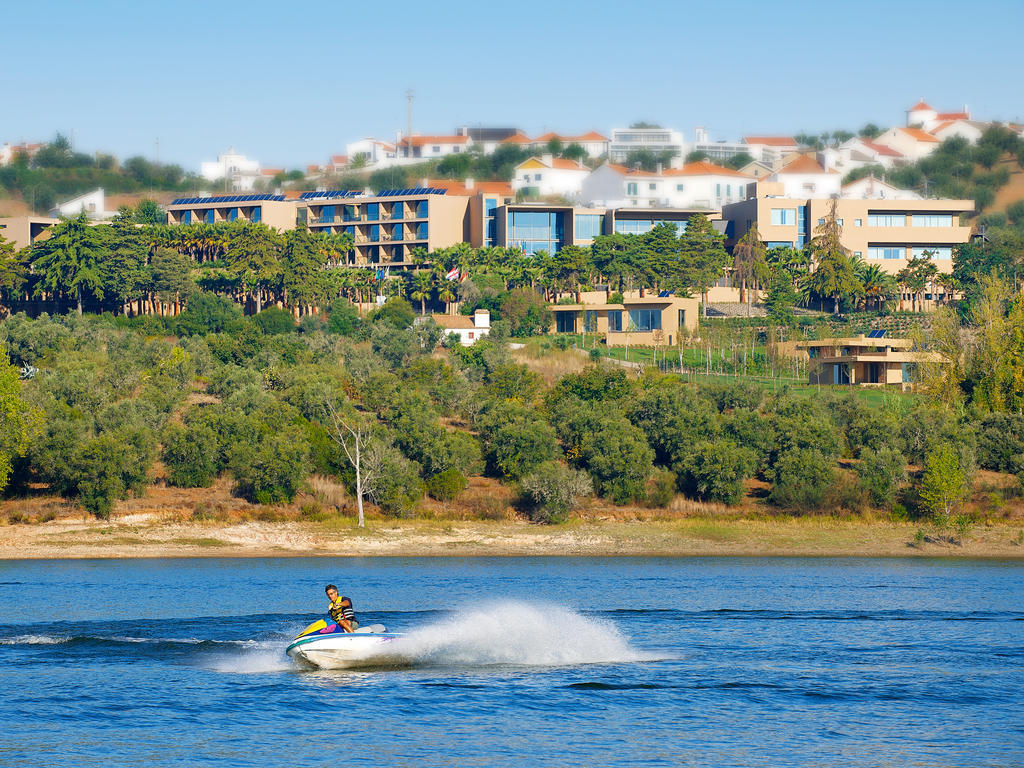Ap Lago Montargil - Conference & Spa Ngoại thất bức ảnh The photo depicts a person riding a jet ski on a body of water. In the background, there are modern buildings and lush greenery, suggesting a scenic landscape, likely near a resort or upscale residential area. The clear blue sky and calm water enhanc