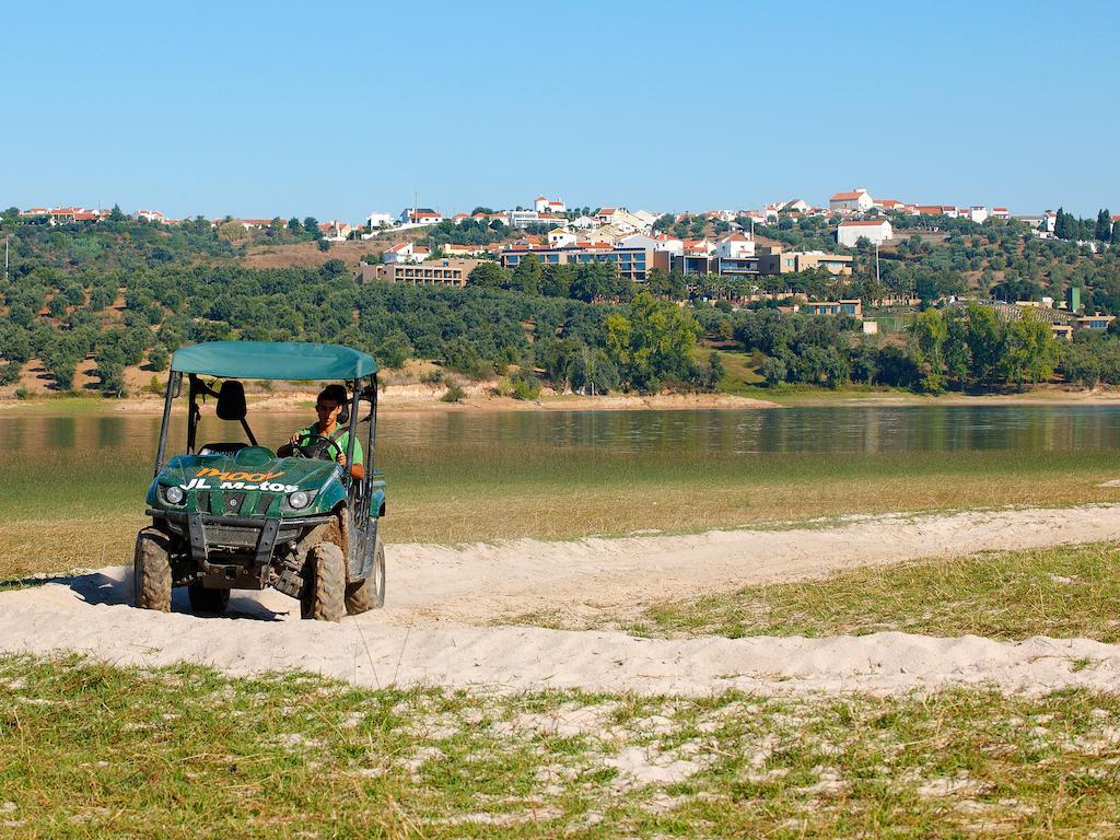 Ap Lago Montargil - Conference & Spa Ngoại thất bức ảnh The photo shows a small green all-terrain vehicle driving along a sandy path beside a body of water. In the background, there's a landscape with trees and rolling hills, along with a town or village visible on the hillside. The sky is clear, indicati