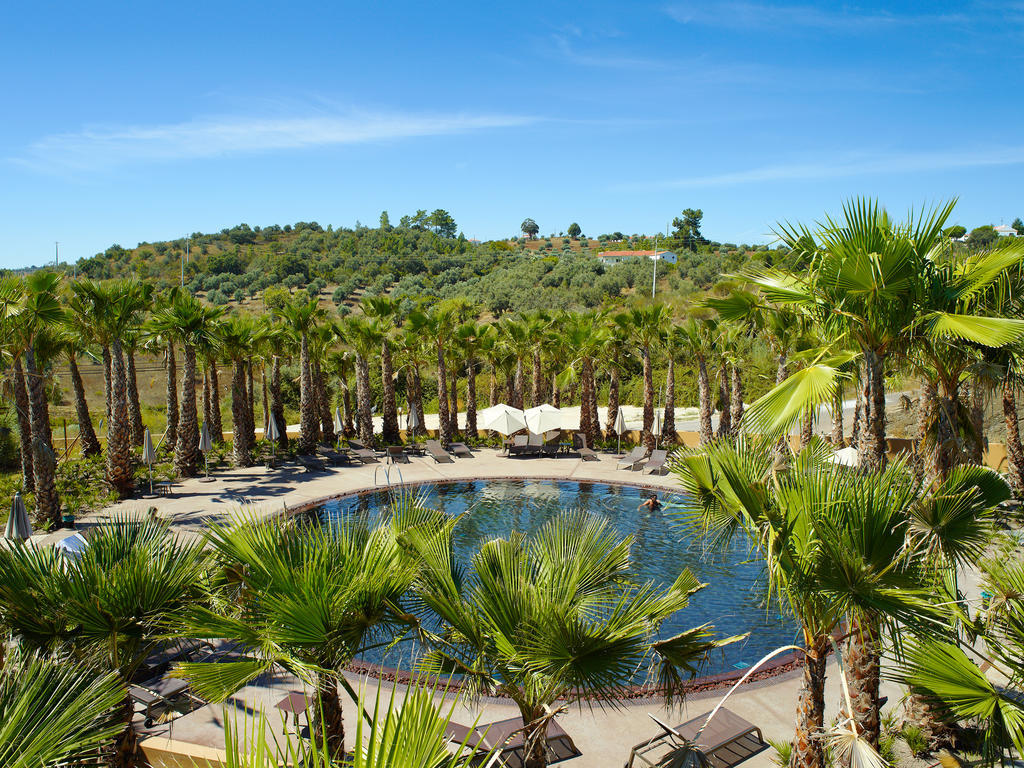 Ap Lago Montargil - Conference & Spa Ngoại thất bức ảnh The photo depicts a beautiful outdoor area featuring a circular swimming pool surrounded by palm trees. The pool area has several lounge chairs arranged around it. In the background, there is a lush green hillside, and the sky is clear with a bright 