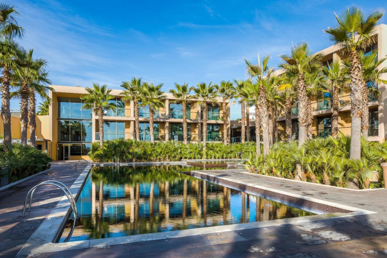 Ap Lago Montargil - Conference & Spa Ngoại thất bức ảnh The photo features a modern building surrounded by lush greenery and palm trees. In the foreground, there is a serene swimming pool with a reflective surface that captures the surrounding environment. The architecture of the building appears to have 
