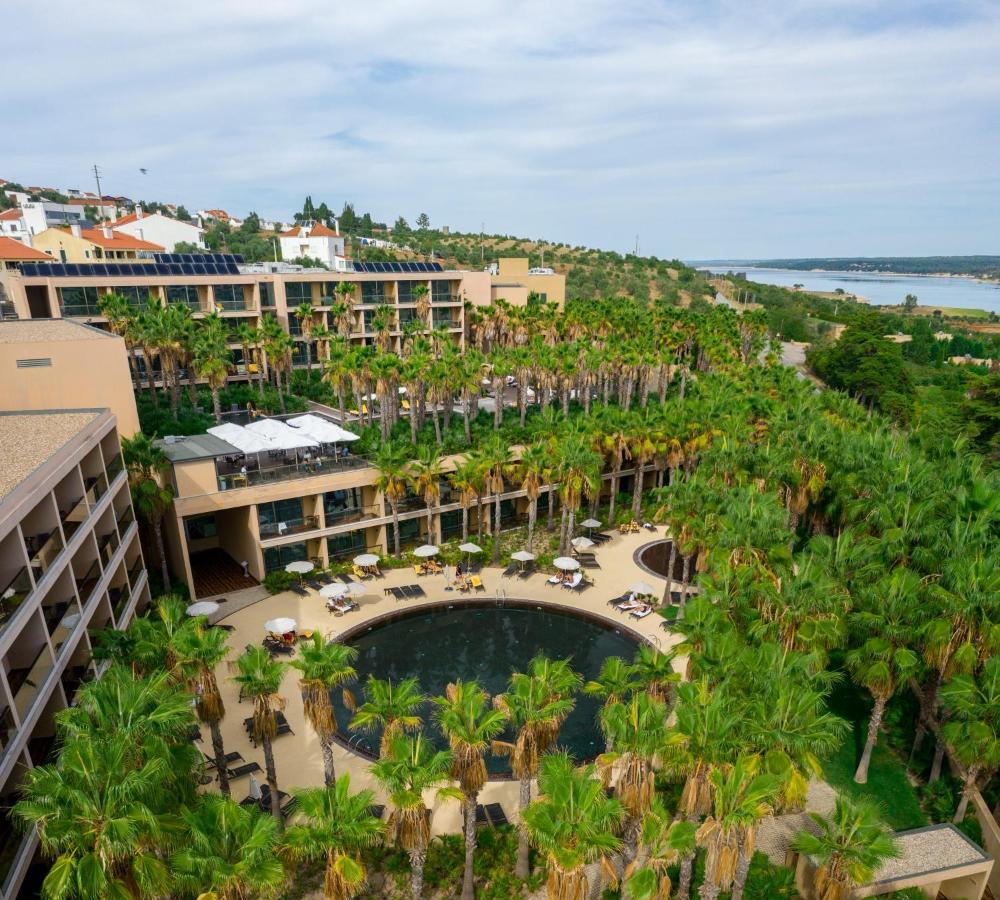 Ap Lago Montargil - Conference & Spa Ngoại thất bức ảnh The photo shows a modern hotel complex set amidst lush greenery, featuring numerous palm trees. In the foreground, there is a circular swimming pool surrounded by lounge chairs. The hotel itself has multiple levels with balconies overlooking the pool