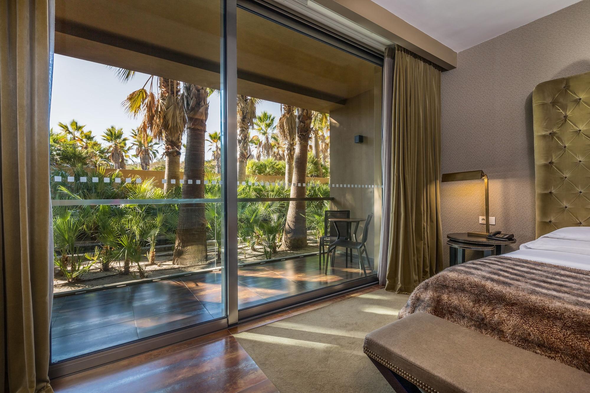 Ap Lago Montargil - Conference & Spa Ngoại thất bức ảnh The photo shows a bedroom with large glass doors that lead out to a balcony or terrace. The view outside features palm trees and lush greenery, indicating a tropical or warm climate. Inside the room, there is a bed, a bedside table with a lamp, and a