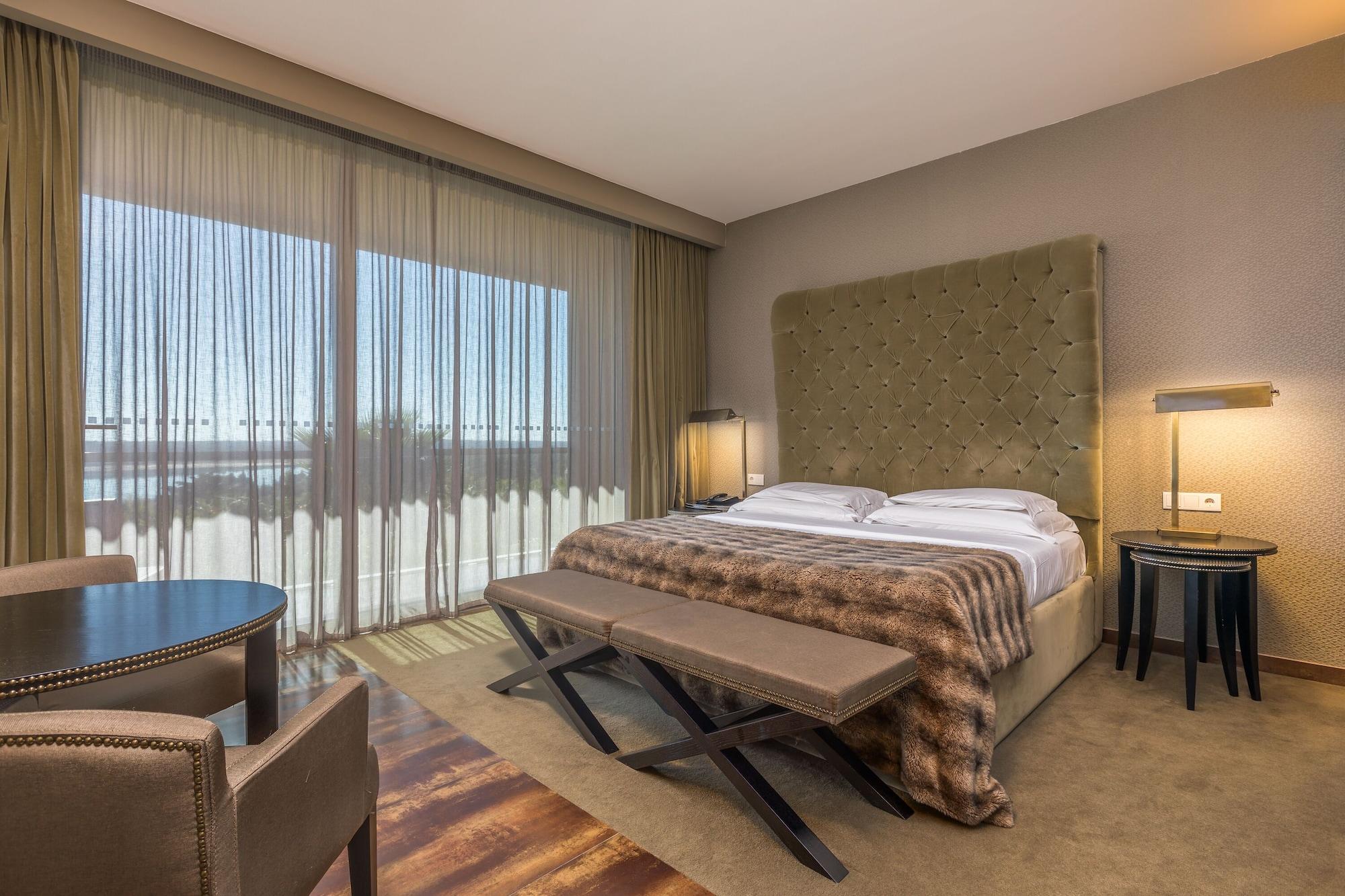 Ap Lago Montargil - Conference & Spa Ngoại thất bức ảnh The photo shows a modern hotel room featuring a large bed with a tufted headboard and a cozy blanket. There are curtains draped over large windows that allow natural light to flow in, providing a view of the outside. A small seating area includes two
