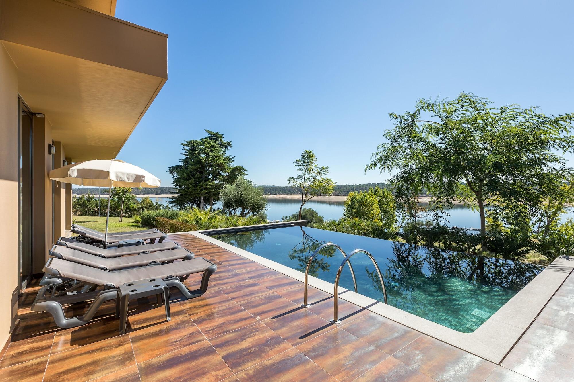 Ap Lago Montargil - Conference & Spa Ngoại thất bức ảnh The photo shows a luxurious outdoor setting by a pool. There are several lounge chairs arranged next to an infinity pool, which overlooks a scenic body of water and lush greenery. A parasol provides shade for relaxation. The sky is clear and blue, in