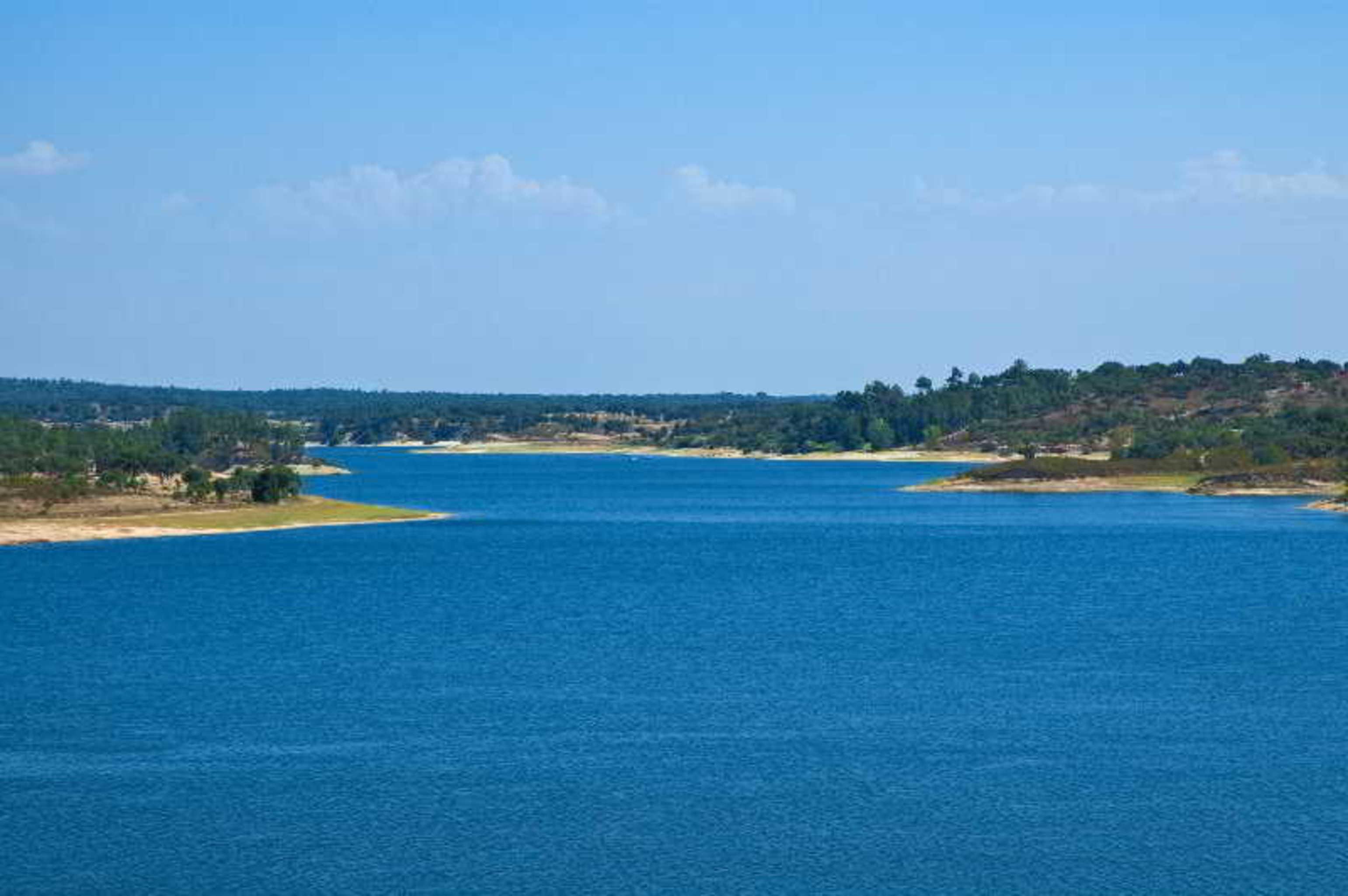 Ap Lago Montargil - Conference & Spa Ngoại thất bức ảnh The photo depicts a serene landscape featuring a large body of water, likely a lake or river. The water appears calm and is reflecting a clear blue sky with some clouds. Surrounding the water, there are patches of green land and trees, indicating a n