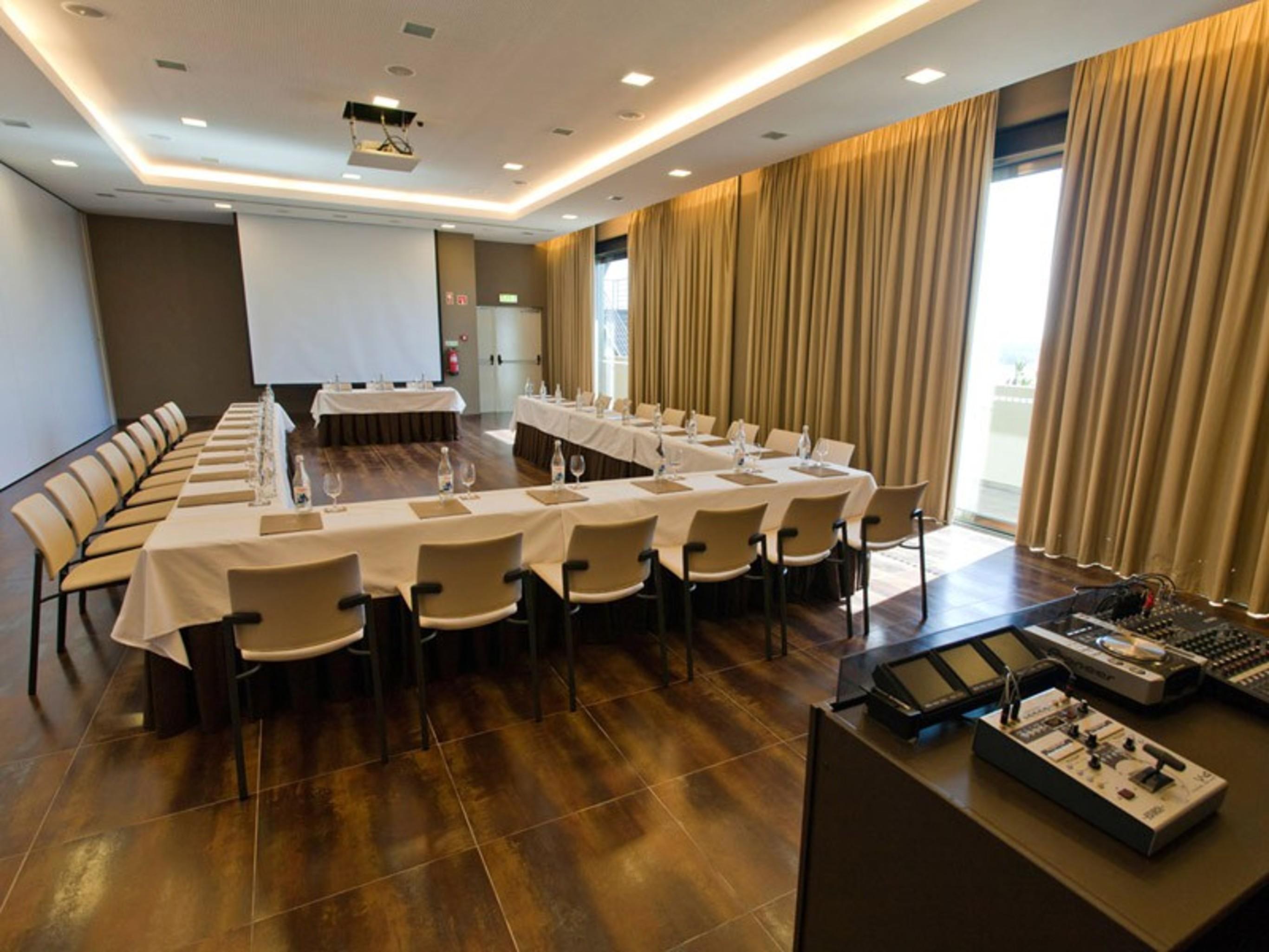 Ap Lago Montargil - Conference & Spa Ngoại thất bức ảnh The photo shows a modern conference room set up for a meeting. There are several tables arranged in a U-shape, surrounded by chairs. A large screen and projector are positioned at the front, with a table for speakers or presenters. The room has large