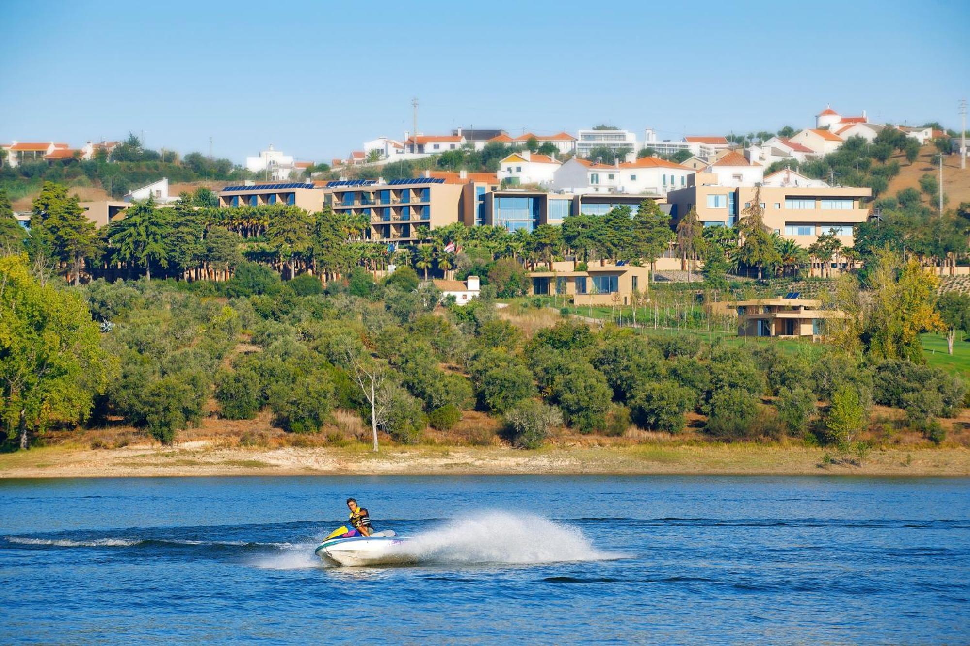 Ap Lago Montargil - Conference & Spa Ngoại thất bức ảnh The photo shows a person riding a jet ski on a body of water, creating a spray as they speed across the surface. In the background, there are modern buildings and a hillside with trees and greenery. The sky is clear and blue, suggesting a bright and 