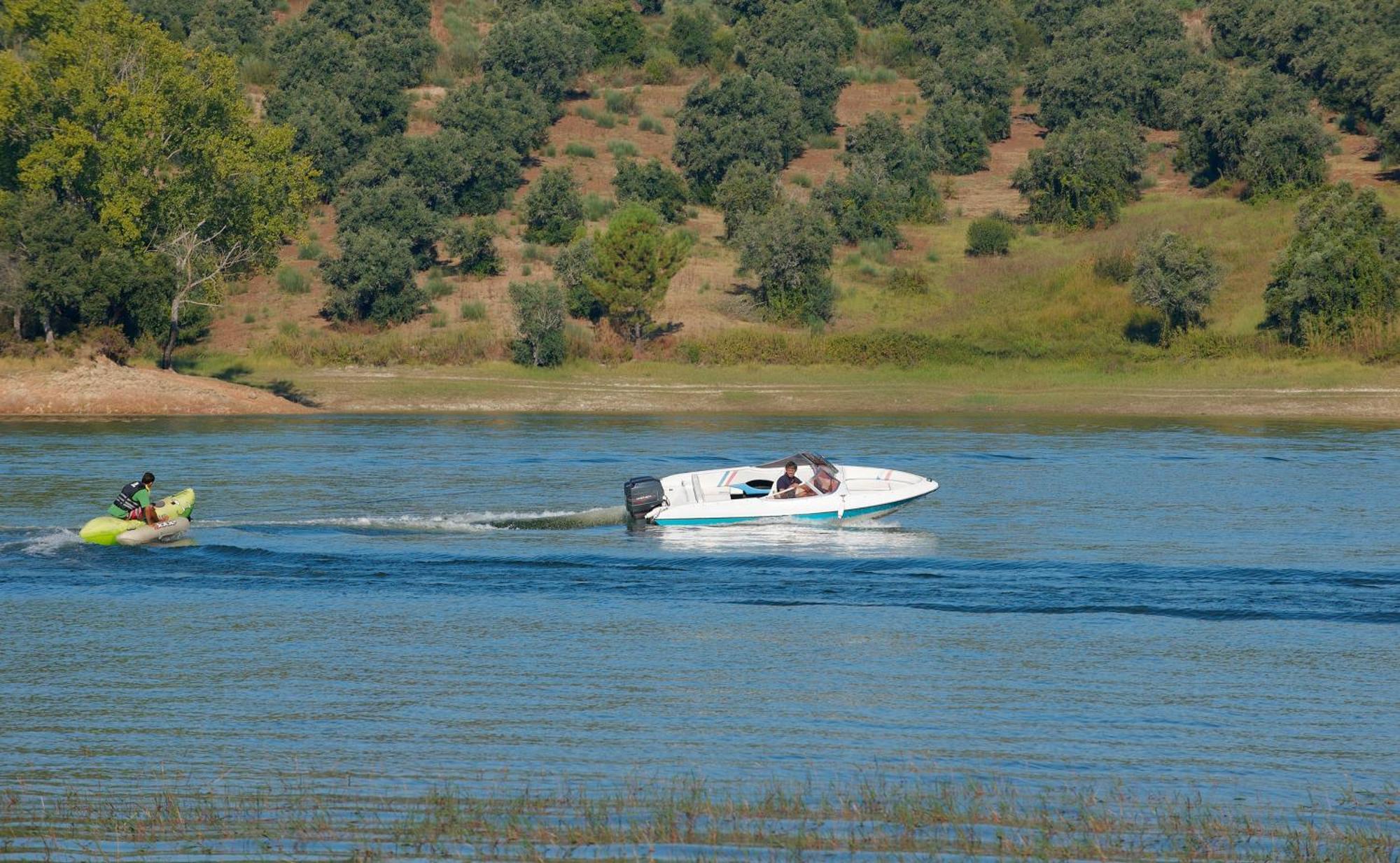Ap Lago Montargil - Conference & Spa Ngoại thất bức ảnh The photo shows a scene on a water body, likely a river or lake. In the foreground, a speedboat is moving quickly through the water, leaving a wake behind it. There are two people visible in the boat. In the background, there is a hilly area covered 