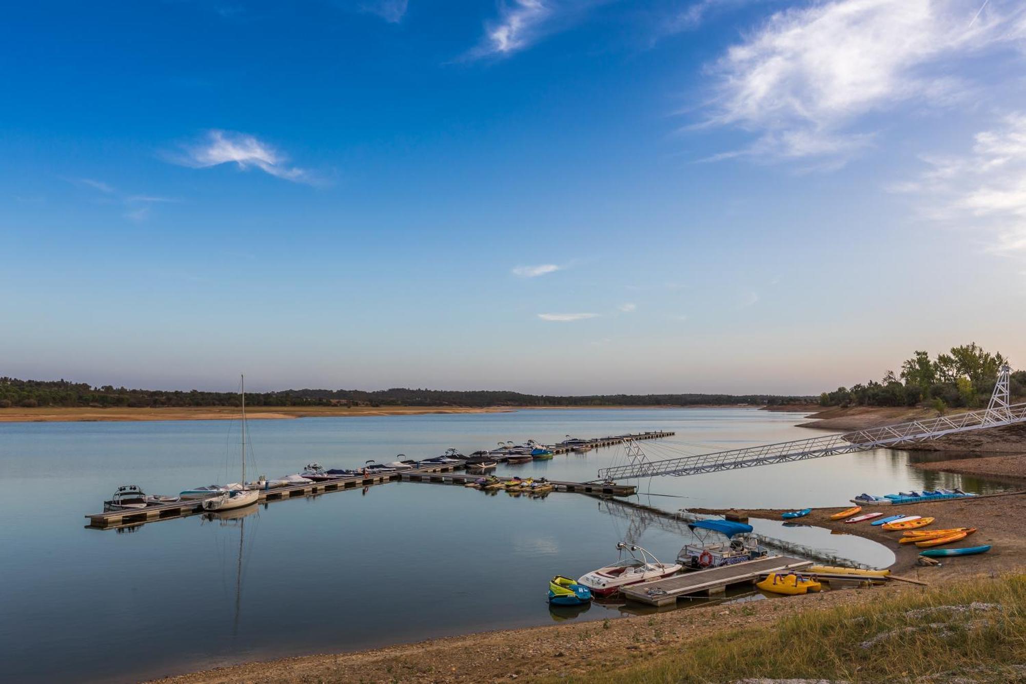 Ap Lago Montargil - Conference & Spa Ngoại thất bức ảnh The photo shows a serene lakeside scene. In the foreground, there are several boats moored at a dock, with some colorful kayaks and canoes lined up along the shore. The water is calm and reflects the sky, which features a few wispy clouds. In the bac