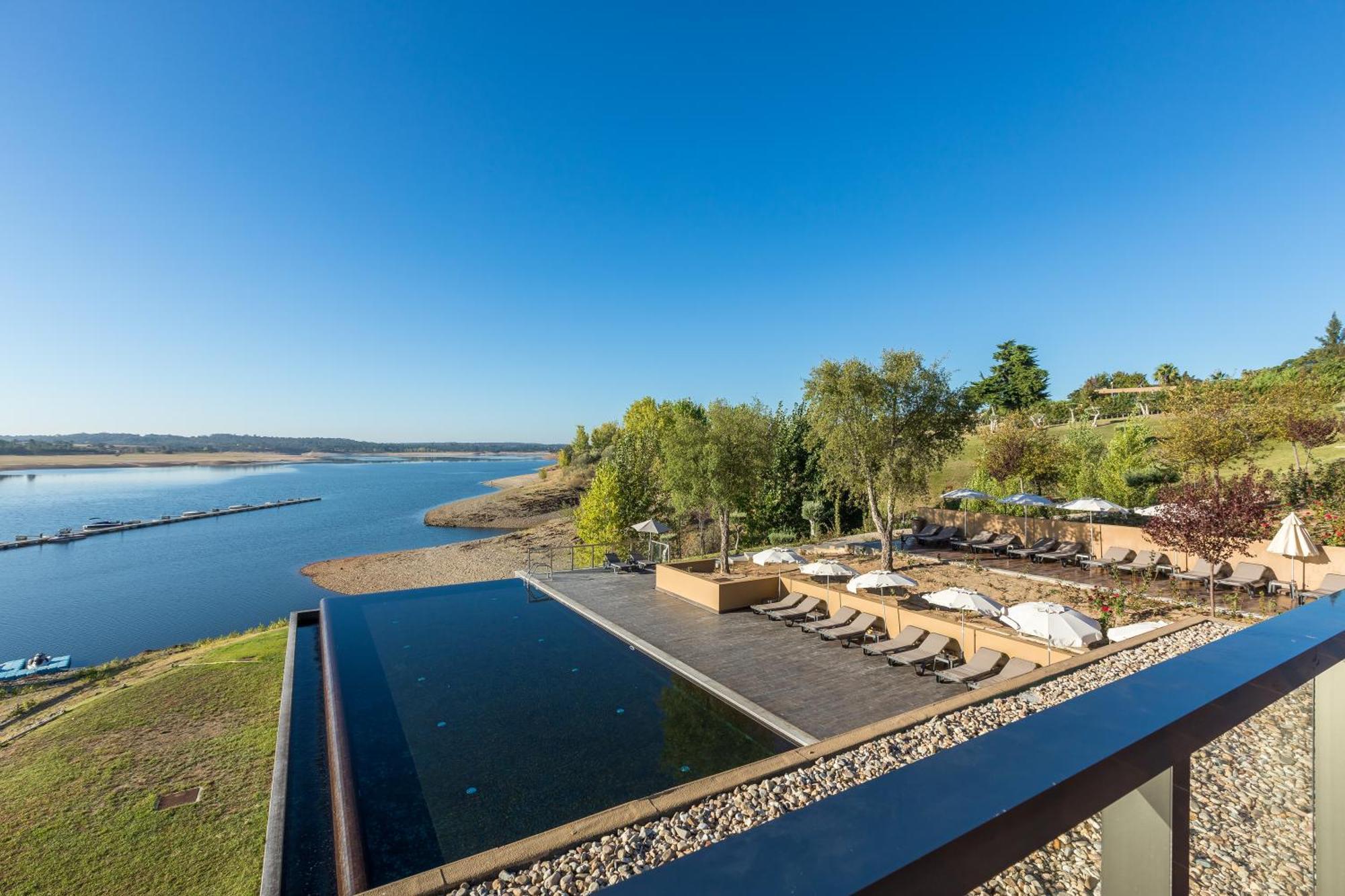 Ap Lago Montargil - Conference & Spa Ngoại thất bức ảnh The photograph depicts a scenic view of a tranquil water body, likely a lake or river, bordered by lush greenery. In the foreground, there is an infinity pool that blends seamlessly with the water beyond. Comfortable lounge chairs are arranged on a w